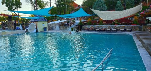 A serene swimming pool surrounded by colorful shade sails and lounge chairs, set in a landscaped area.