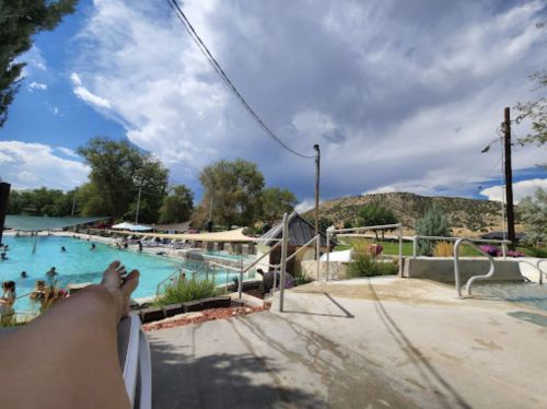 A pool scene with people swimming, surrounded by trees and mountains under a partly cloudy sky. A foot is visible in the foreground.