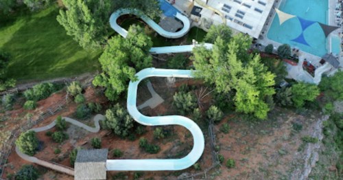 Aerial view of a winding water slide surrounded by trees, with a pool and building in the background.