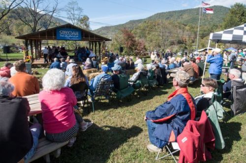 A crowd gathers at an outdoor event with a stage, surrounded by mountains and trees on a sunny day.