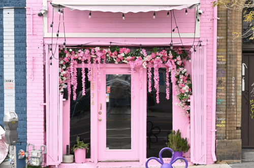 A vibrant pink storefront adorned with flowers and greenery, featuring a large glass door and decorative lights.