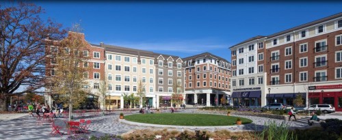 A vibrant urban plaza with green space, surrounded by modern buildings and shops under a clear blue sky.