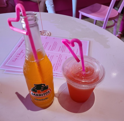 Two drinks on a table: a bottle of Jarritos soda and a cup with a colorful straw, set in a pink-themed café.