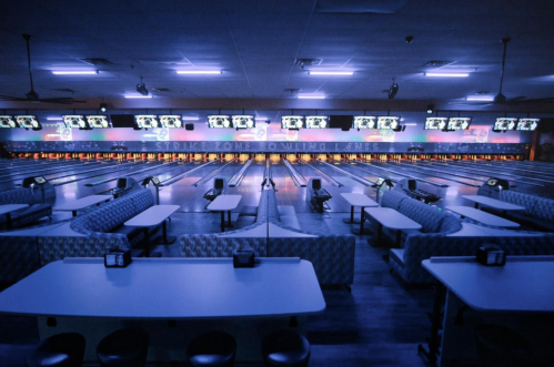 A dimly lit bowling alley with empty lanes, blue lighting, and seating areas. Screens display scores above the lanes.