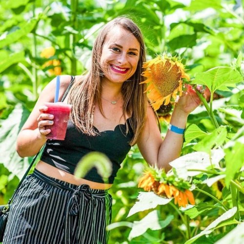 A smiling woman holds a sunflower and a drink while standing among tall sunflowers in a bright, sunny field.