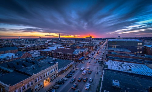 Aerial view of a city at sunset, with vibrant clouds and busy streets lined with cars and buildings.