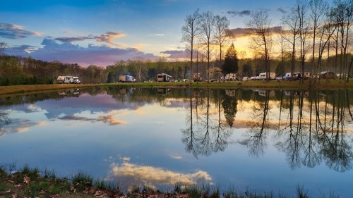 A serene lake reflects trees and clouds at sunset, with campers visible along the shore.
