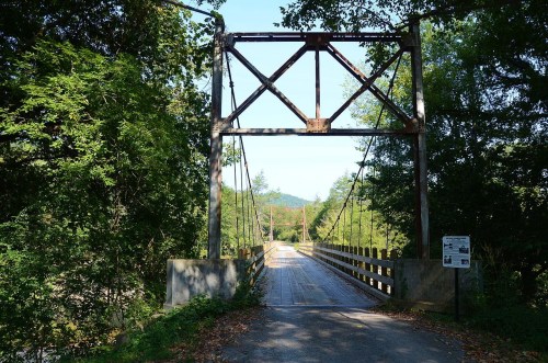 A metal truss bridge spans a river, surrounded by lush greenery and trees under a clear blue sky.