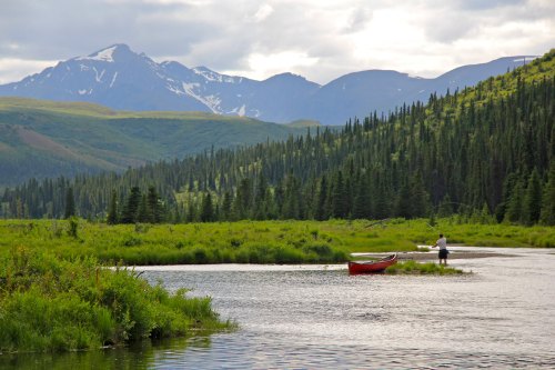 A person stands in a red canoe on a calm river, surrounded by lush greenery and mountains in the background.