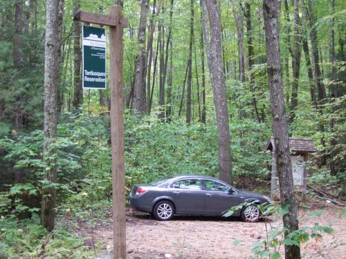 A gray car parked near a sign for Tantasqua Reservation, surrounded by lush green trees and foliage.