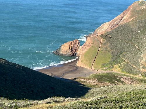 A scenic view of a rocky coastline with a sandy beach, surrounded by green hills and blue ocean waves.