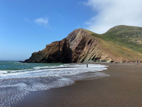 A person stands on a sandy beach near a rocky cliff, with gentle waves lapping at the shore under a clear blue sky.