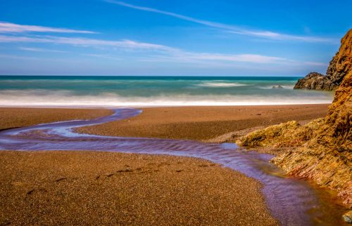 A serene beach scene with gentle waves, a winding stream, and rocky cliffs under a clear blue sky.