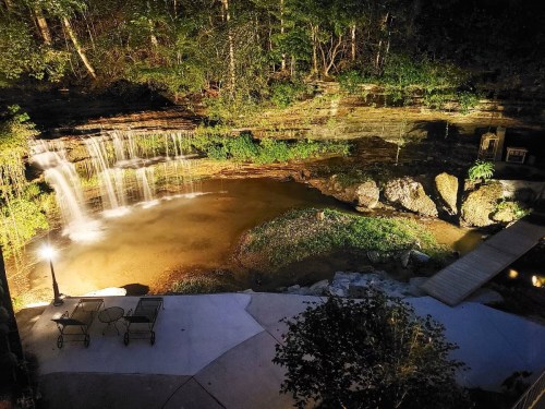A serene nighttime scene of a waterfall cascading into a pond, surrounded by lush greenery and soft lighting.