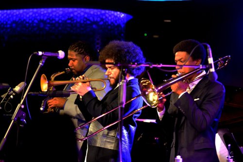 Three musicians play trombones on stage, illuminated by colorful lights, creating a lively jazz atmosphere.