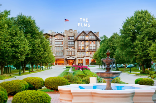 Historic hotel "The Elms" with a fountain in front, surrounded by lush greenery and a clear blue sky.