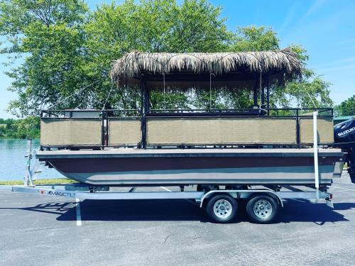 A boat with a thatched roof on a trailer, parked by a lake surrounded by trees.