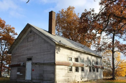 A weathered wooden house with a chimney, surrounded by autumn trees and a clear blue sky.