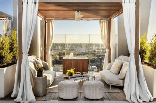 A stylish outdoor lounge with white curtains, comfortable seating, and a view of the city skyline.