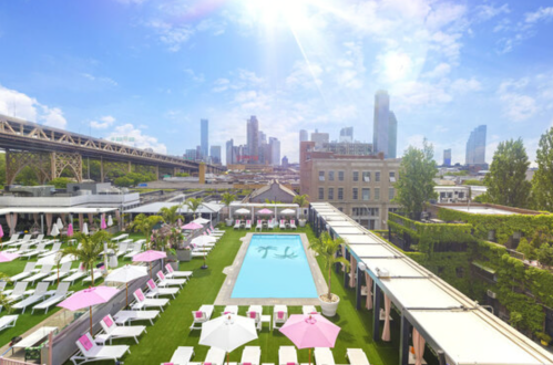 A rooftop pool area with lounge chairs, palm trees, and a city skyline under a bright blue sky.