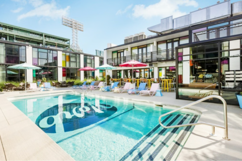 A vibrant pool area with colorful umbrellas and lounge chairs, surrounded by modern buildings and a clear blue sky.
