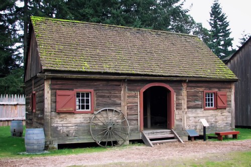 A rustic wooden cabin with a mossy roof, red shutters, and a wagon wheel, surrounded by trees and grass.