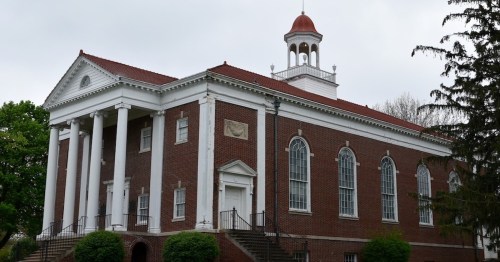 Historic brick building with white columns, a red roof, and a bell tower, surrounded by greenery under a cloudy sky.