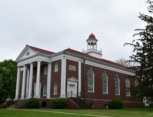 A brick building with white columns, a red roof, and a cupola, surrounded by greenery under a cloudy sky.