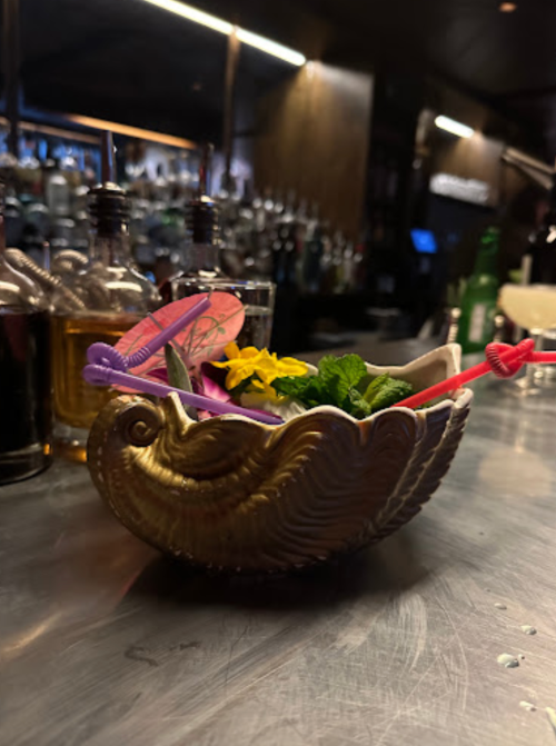 A decorative bowl filled with colorful garnishes, including flowers and mint, sits on a bar counter.