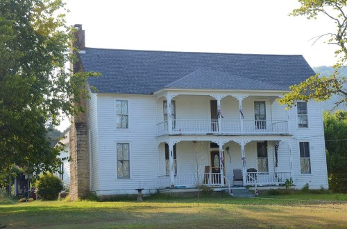 A two-story white house with a porch, surrounded by greenery and trees, set against a hillside backdrop.