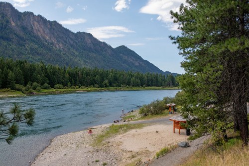 A serene river scene with mountains in the background, surrounded by lush trees and a sandy beach area.