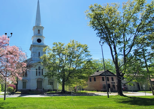A tall church with a spire surrounded by trees and a pink flowering tree, next to an old brick building on a sunny day.