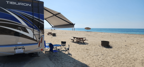 A camper parked on a sandy beach with picnic tables and an umbrella, overlooking the ocean under a clear blue sky.