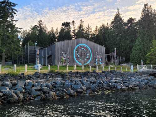 A wooden building with a large mural, surrounded by trees and rocky shoreline, under a cloudy sky.