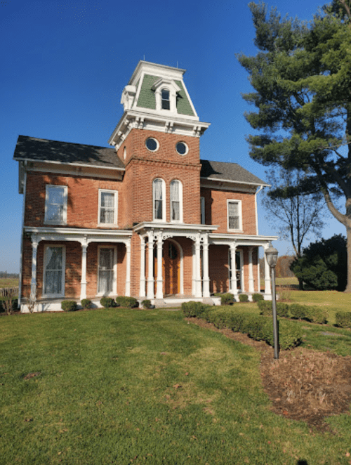 A historic brick house with a green roof, white columns, and a tower, surrounded by a well-maintained lawn and trees.