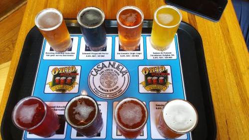 A tray with eight different beer samples in small glasses, arranged on a branded mat with brewery information.