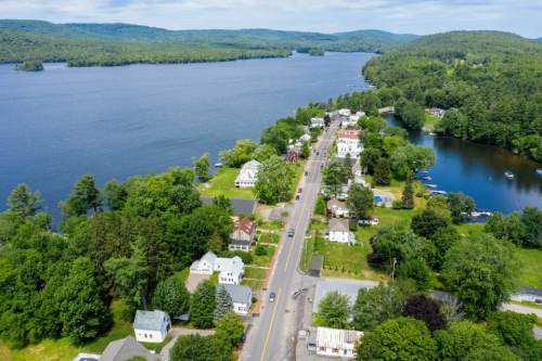 Aerial view of a serene lakeside town with green trees, houses, and a winding road along the water's edge.