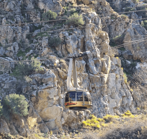 A gondola glides over rocky terrain, suspended by cables against a backdrop of rugged cliffs and greenery.