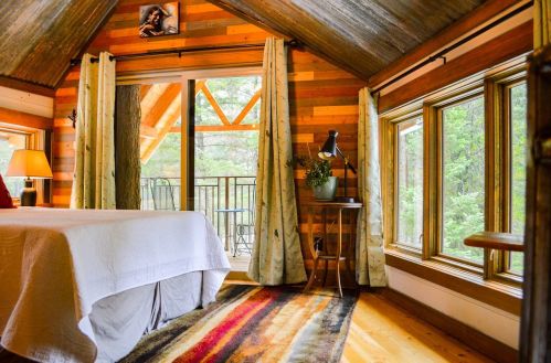 Cozy cabin interior with wooden walls, a bed, large windows, and a balcony overlooking nature. Warm, inviting atmosphere.
