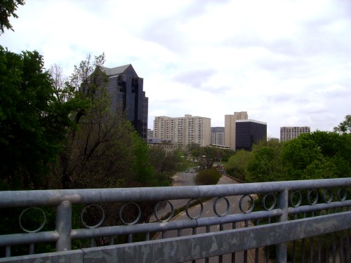 View from a bridge overlooking a city skyline with modern buildings and greenery under a cloudy sky.