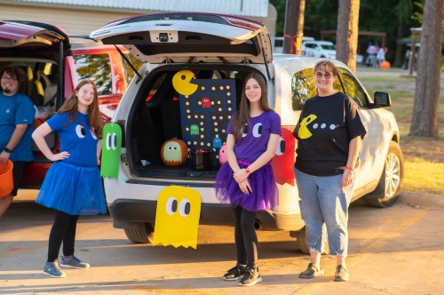 Two girls in colorful outfits pose next to a decorated car trunk featuring a Pac-Man theme, with a woman beside them.