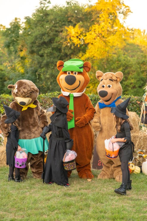 Two costumed characters in bear suits and two children in witch costumes gather outdoors with Halloween treats.