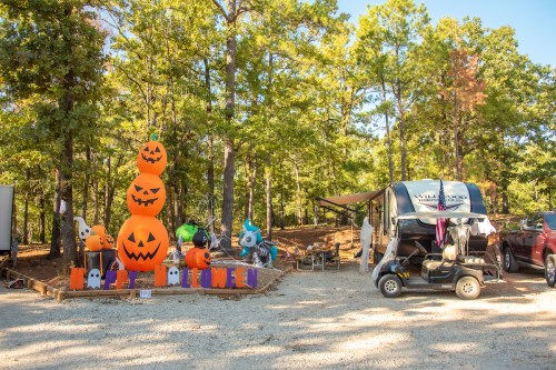 A festive outdoor scene with stacked pumpkin decorations, ghosts, and a golf cart in a wooded area.