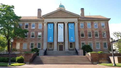 Historic building with columns and large banners displaying "UNC," surrounded by greenery and a clear blue sky.