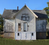 A weathered, two-story house with a white facade and a grassy foreground under a clear blue sky.