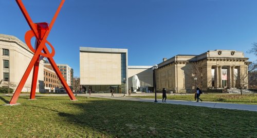 A modern building and a classical structure are seen beside a large red sculpture on a sunny day.