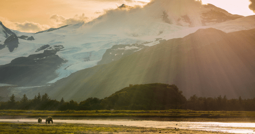 Two bears wander along a riverbank with majestic mountains and sunlight streaming through clouds in the background.