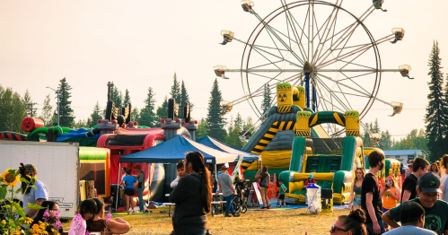 A lively fair scene with a ferris wheel, inflatable attractions, and people enjoying the festivities under a clear sky.