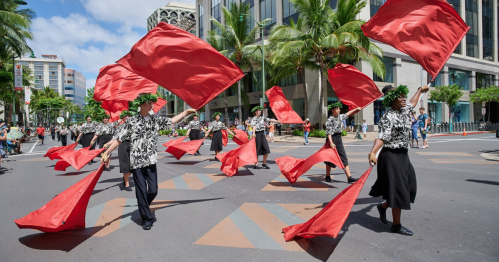 A vibrant parade scene with dancers in floral shirts waving red flags amidst palm trees and a city backdrop.
