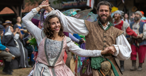 A man and woman in historical costumes dance joyfully at a festival, surrounded by a lively crowd.
